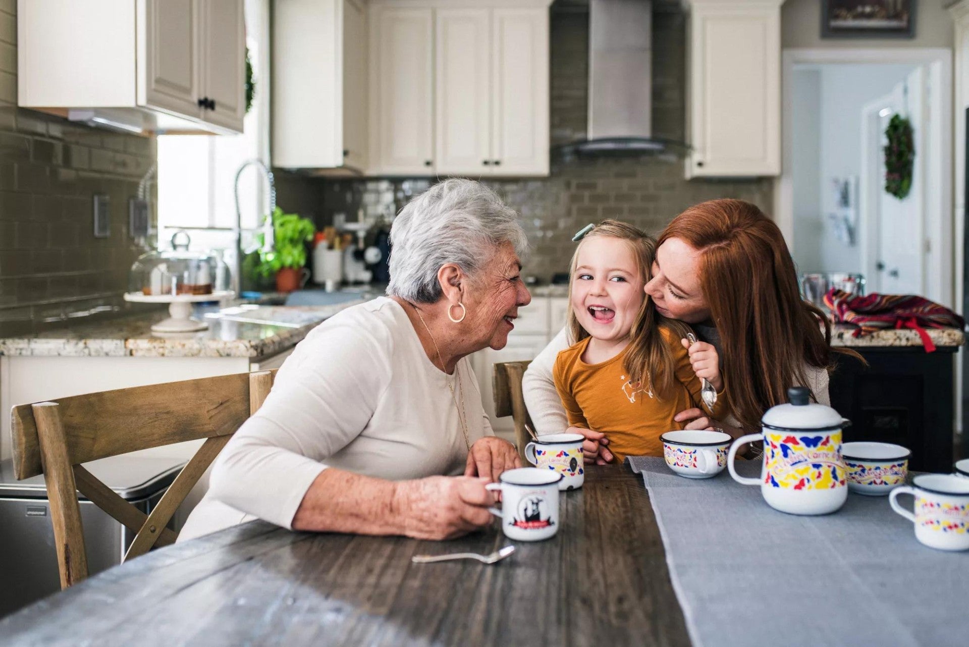 Mother kissing daughter on the cheek at kitchen table with grandma laughing. Mother kissing daughter on the cheek at kitchen table with grandma laughing.