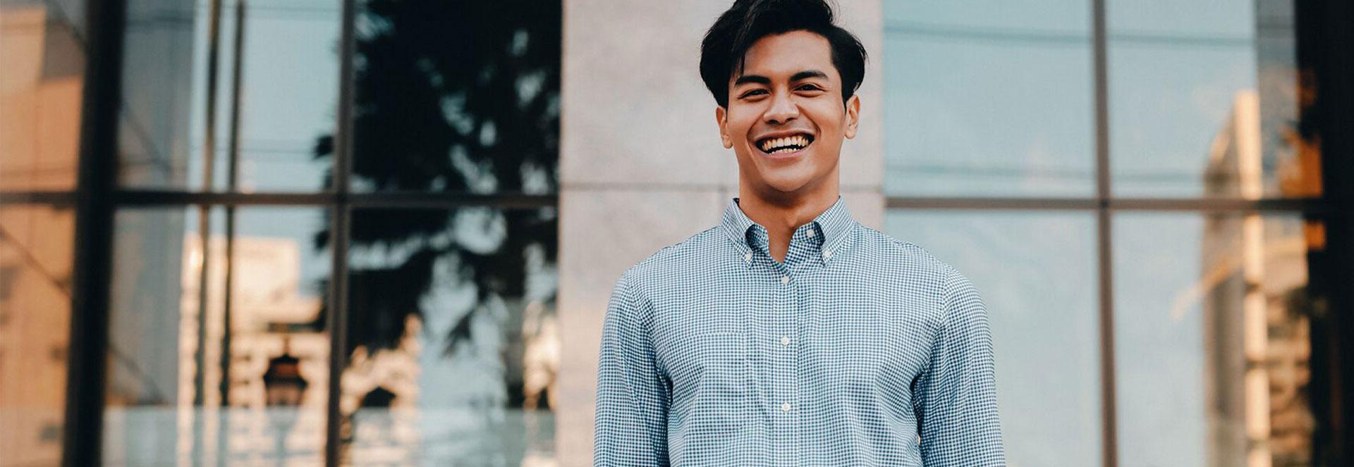 Young man in blue button-up shirt standing in front of an office building with a big smile