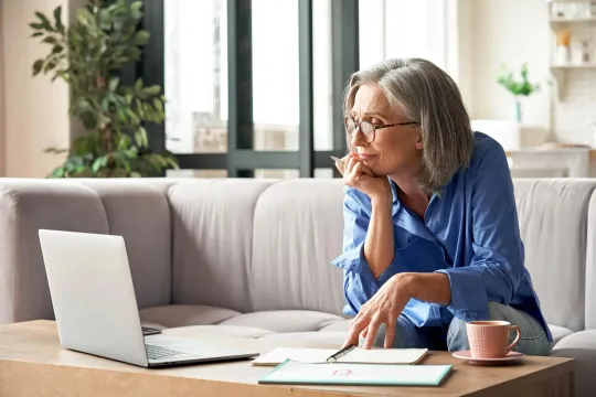 Woman in her living room resting her chin on her hand as she looks at her laptop.