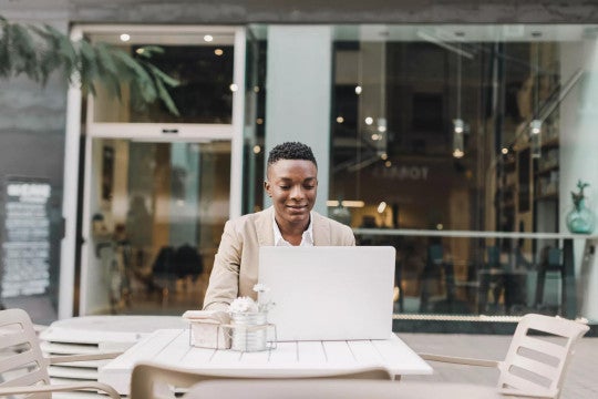 Young man working on his laptop at a patio table outside of a business. 
