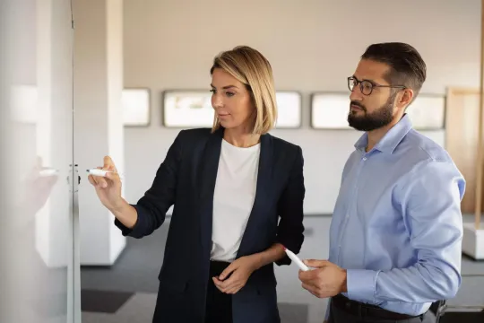 Two employees, a woman and a man, looking at and writing on a whiteboard in the office. 