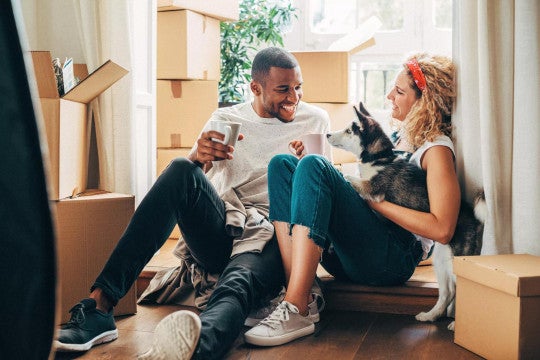 Man and woman sit on floor of home holding dog and surrounded by moving boxes.
