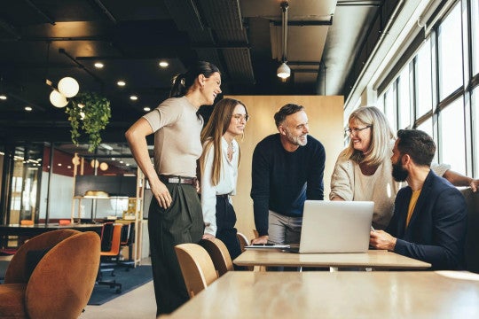 Five colleagues of various ages working together around a laptop in a modern office environment.