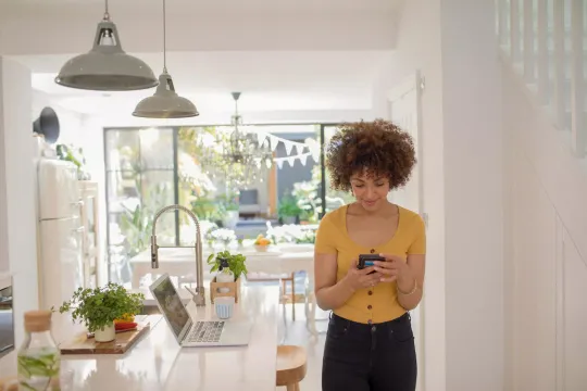 Young woman using smart phone and computer to check health savings account standing in sunny kitchen.