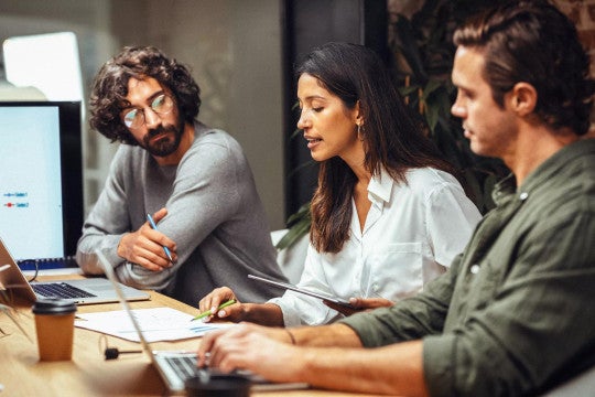 Three employees collaborating at a desk looking at their computers and documents.