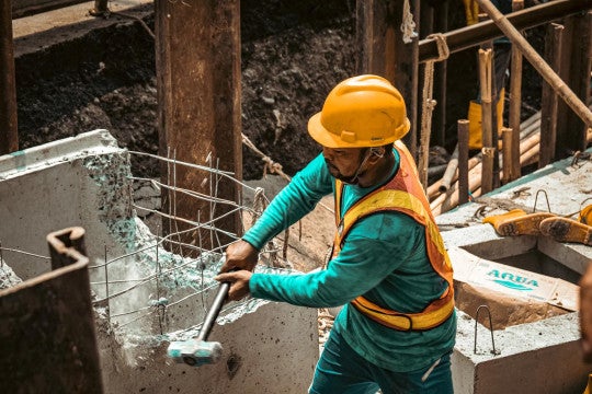 Worker in a hardhat and orange safety vest using a sledgehammer to break up a piece of concrete.