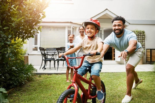 Dos padres con sus dos niños jugando en el patio, uno de ellos aprendiendo a montar en bicicleta.