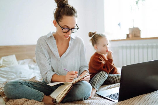 Mother and her young daughter sitting on a bed with a notebook in hand and laptop open.