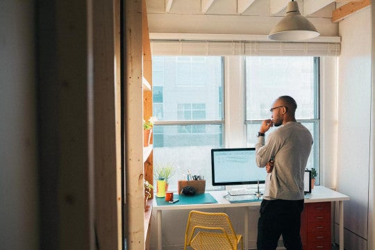A man standing in a modern, sunlit home office, looking thoughtfully at a computer screen.