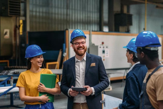 A day in the factory, workers and managers with tablets and clipboards in hard hats smiling in conversation.