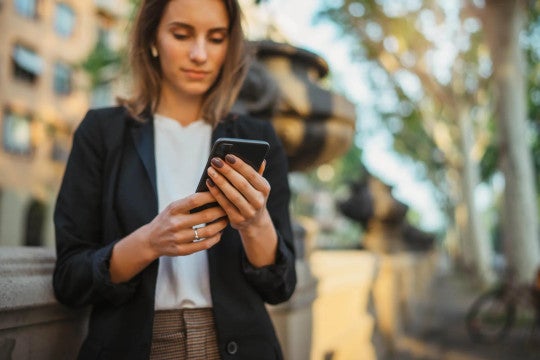 A person in professional attire wearing a black blazer and white top checks their mobile phone while standing outdoors
