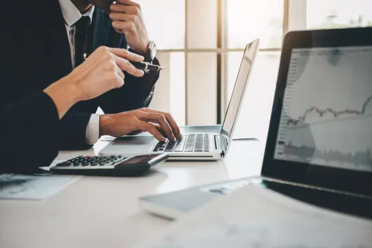 Business professional typing on laptop at desk with financial charts visible, photographed from side angle in natural light.