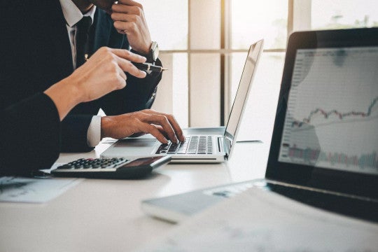 Business professional typing on laptop at desk with financial charts visible, photographed from side angle in natural light.
