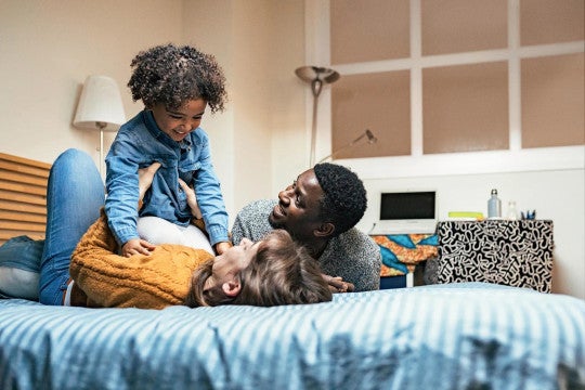 A joyful family moment as parents and child play together on a bed, laughing and being playful in a casual home setting.