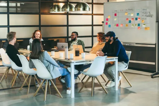 Six employees around a desk having a brainstorming session in front of a whiteboard with sticky notes. 