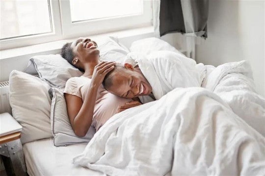 Young couple laughing together while relaxing in bed with white bedding, sharing a joyful morning moment