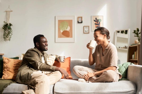 Two people relaxing on a couch, sharing coffee and laughing in a bright living room with modern wall art and plants