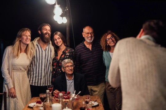 A group of people smiling at an evening gathering around a table with desserts, under string lights. Casual, warm atmosphere.