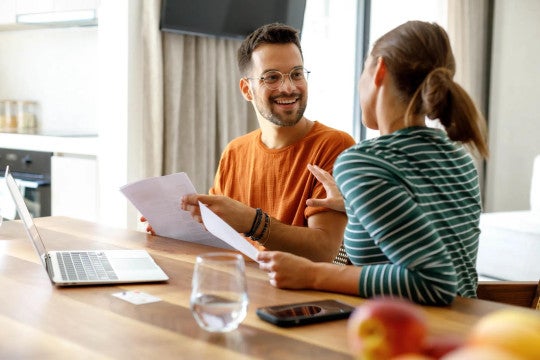 Two colleagues review documents at desk with laptop, one wearing orange shirt and one in striped top, in bright office setting