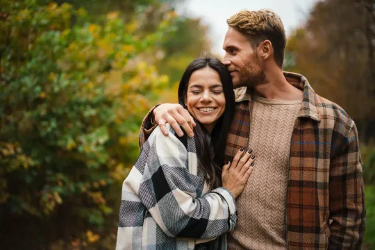 A couple embracing outdoors in fall, wearing cozy plaid and knit sweaters. Autumn foliage in background.