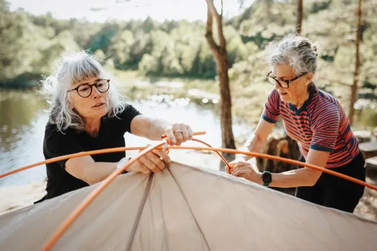 Two retirement age women setting up a tent together beside a lake in a sunny forested camping area.