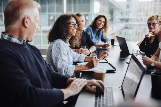 Diverse group of professionals collaborate in bright office setting, sharing ideas over laptops with coffee in a casual meeting.