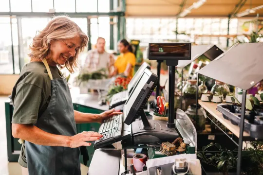 A florist smiles as she operates the cash register in her greenhouse, with customers in the background. 
