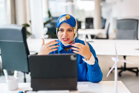 A businesswoman wearing a hijab conducts a videoconference on her laptop computer while sitting in her office. 