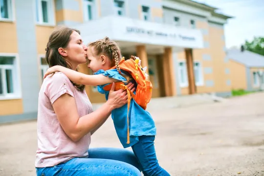 Mom sitting on a curb giving a kiss to daughter wearing a backpack