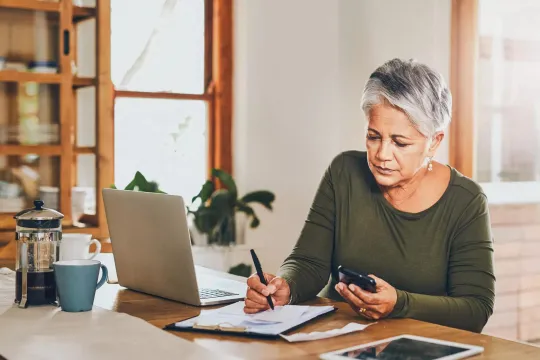 Women working at a table with her laptop and cell phone, writing on paper. 