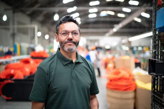 Person wearing a green shirt stands in a warehouse with stacked boxes and coiled orange hoses in the background