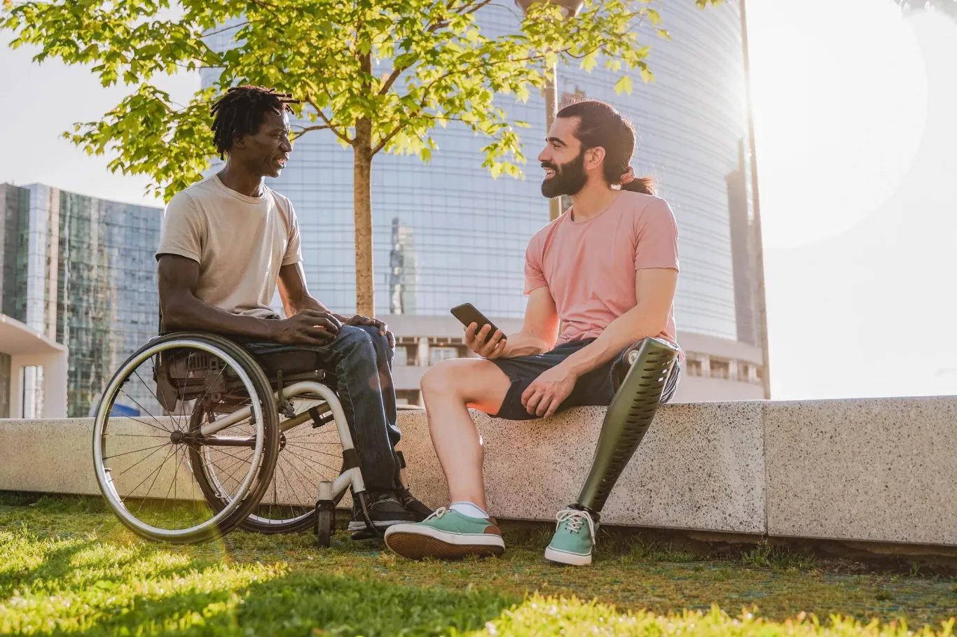 Black man in wheelchair and sitting ethnic man with prosthetic leg conversing in outside courtyard.