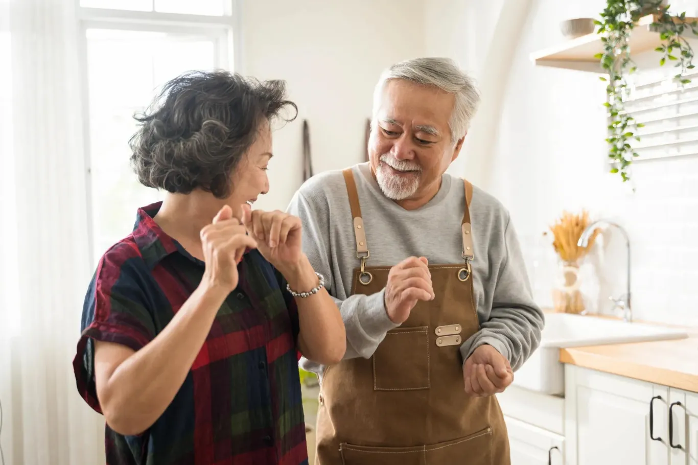 Senior couple dancing together in bright kitchen, man wearing overalls, woman in plaid, both smiling and enjoying moment