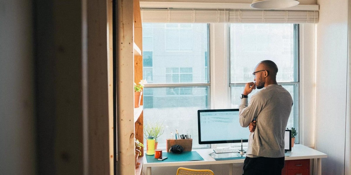 A man standing in a modern, sunlit home office, looking thoughtfully at a computer screen.