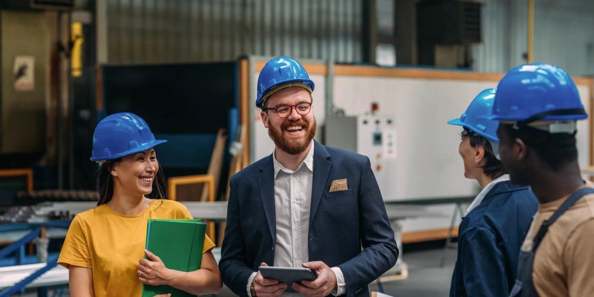 A day in the factory, workers and managers with tablets and clipboards in hard hats smiling in conversation.
