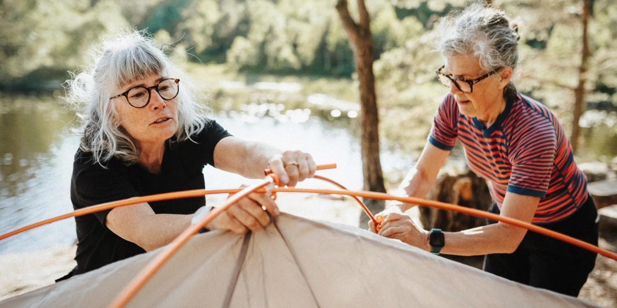 Two retirement age women setting up a tent together beside a lake in a sunny forested camping area.
