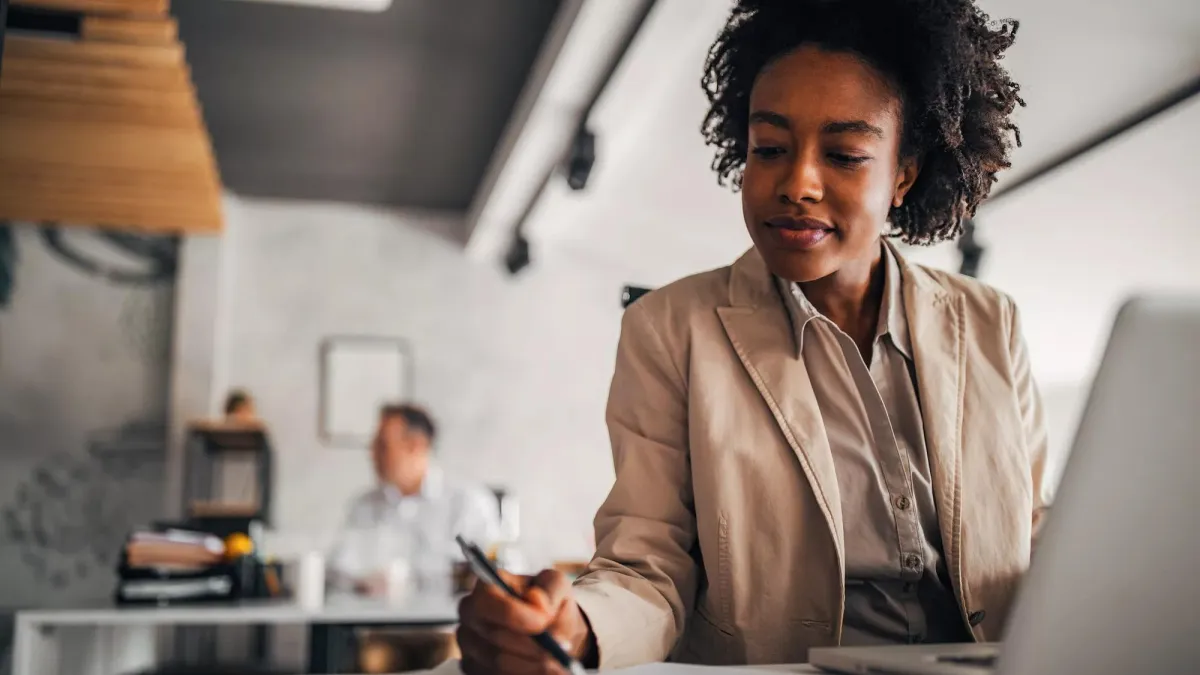 Woman working at laptop also writing a note on a nearby sheet of paper.