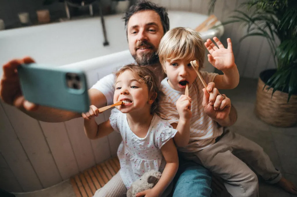 A man taking a selfie with his two young children while they are brushing their teeth.