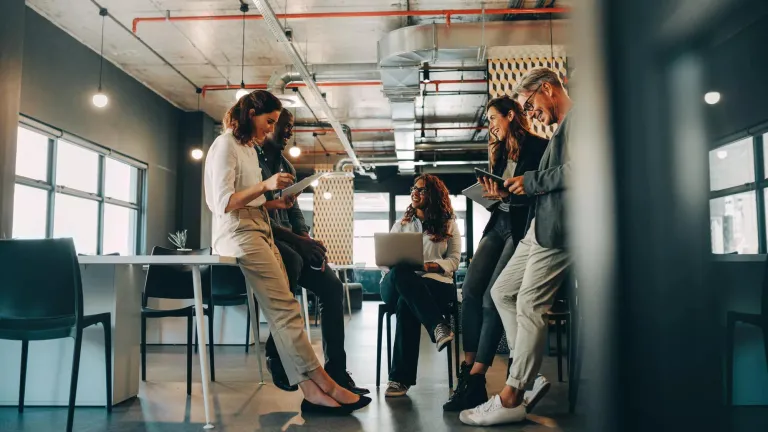 Five young men and women gather in a semi-circle, smiling and collaborating on a work project.