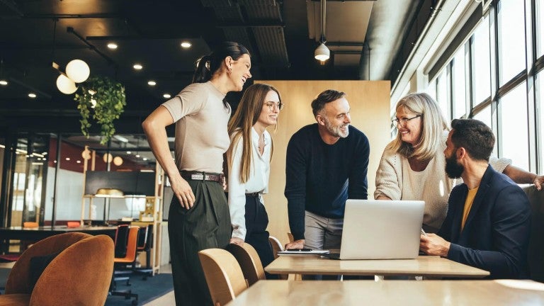 Five colleagues of various ages working together around a laptop in a modern office environment.