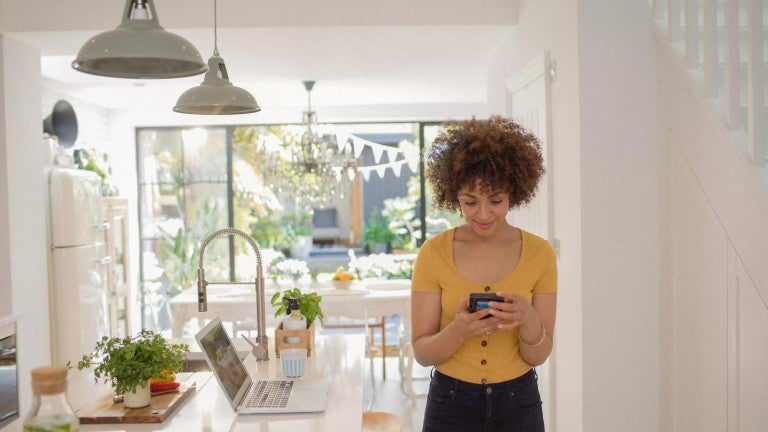 Young woman using smart phone and computer to check health savings account standing in sunny kitchen.