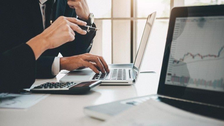 Business professional typing on laptop at desk with financial charts visible, photographed from side angle in natural light.