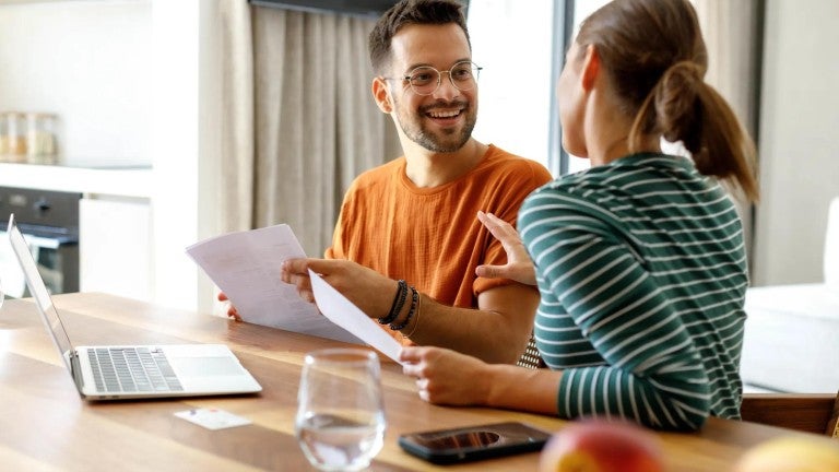 Two colleagues review documents at desk with laptop, one wearing orange shirt and one in striped top, in bright office setting