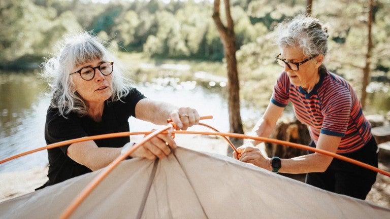 Two retirement age women setting up a tent together beside a lake in a sunny forested camping area.