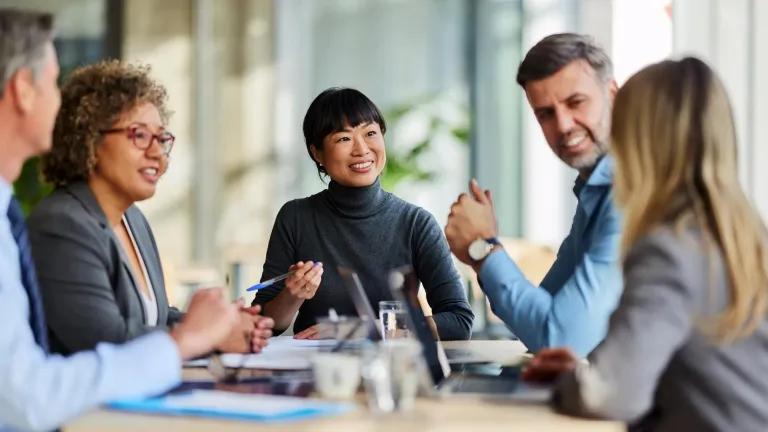 Group of professionals seated around a table in an office, engaged in discussion with documents, pens, and laptops visible.