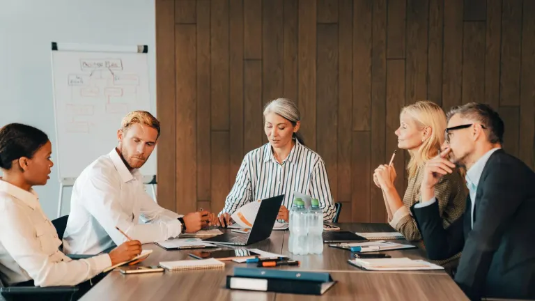 Six professionals seated at a conference table reviewing documents during a meeting.