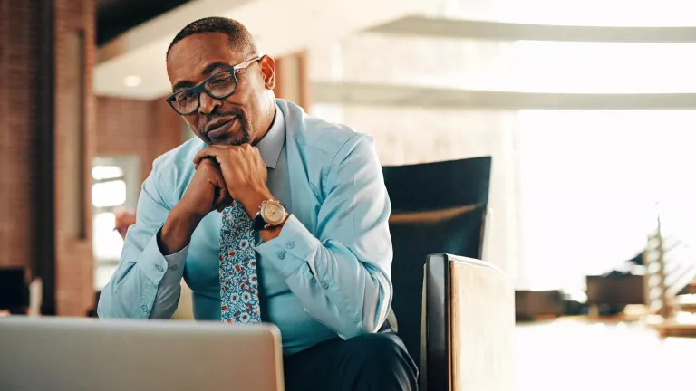 Person sitting in office chair, hands clasped, looking at laptop, wearing glasses, dress shirt, and sassy tie.