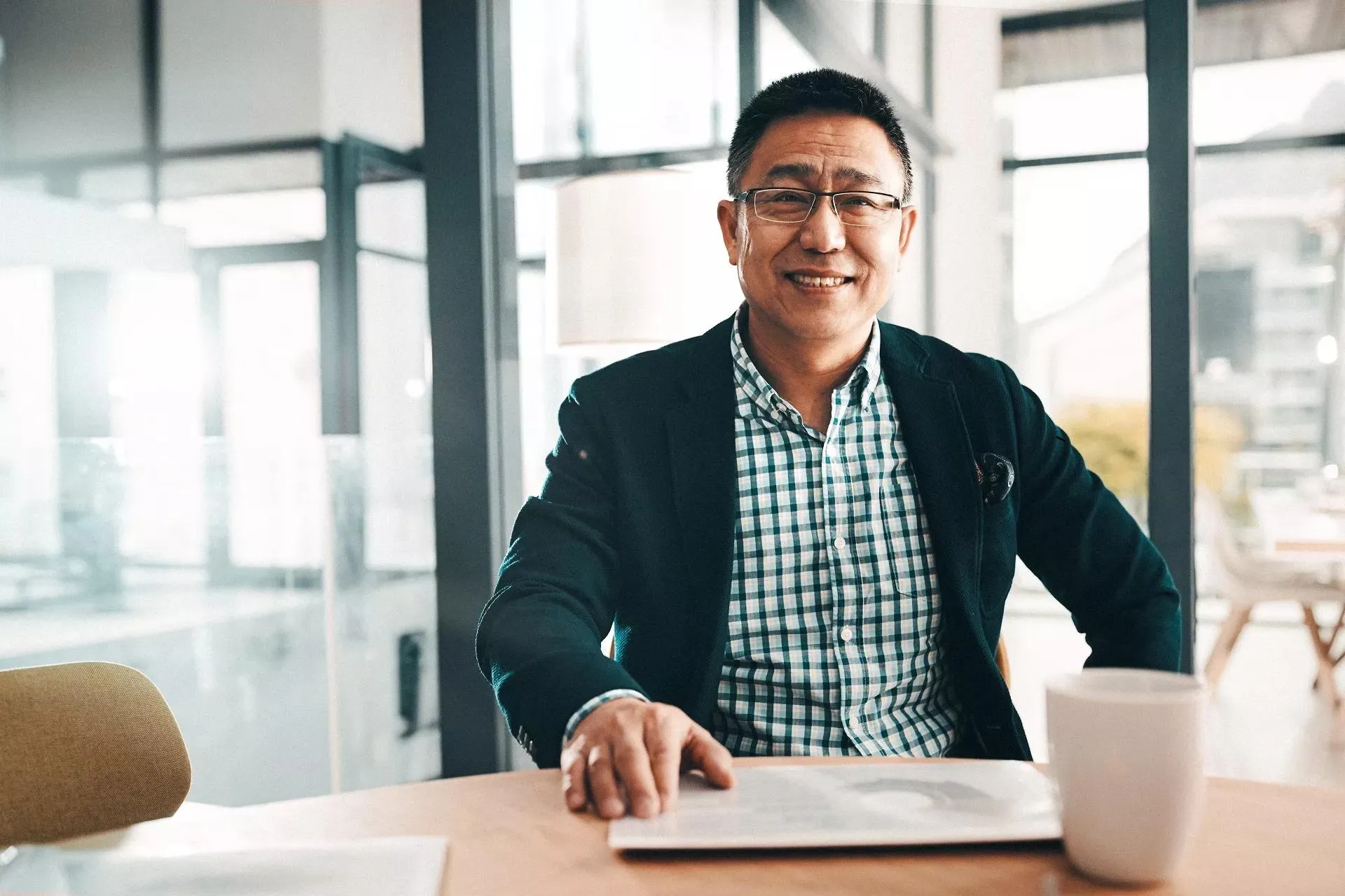 Asian man sitting at a table smiling while drinking coffee and reading a newspaper.