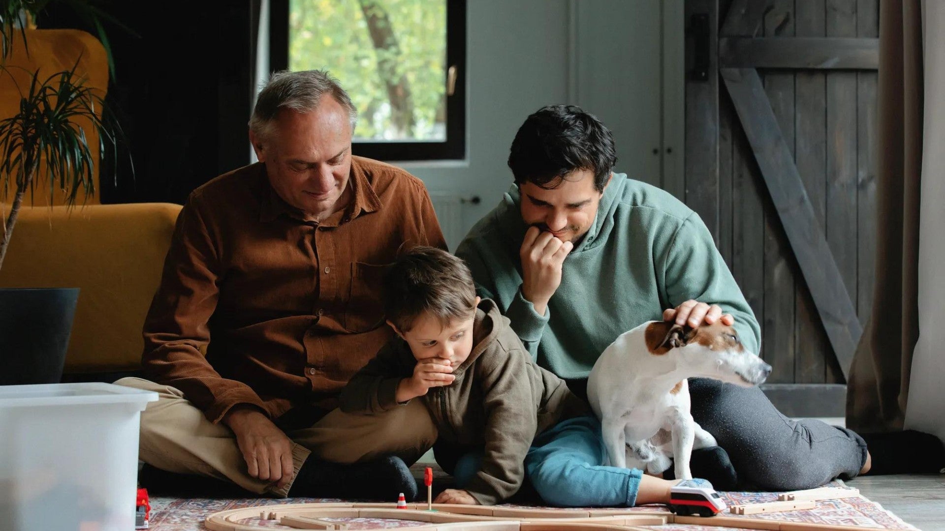 A grandfather, his son, and grandson playing with trains on the living room floor with their dog.