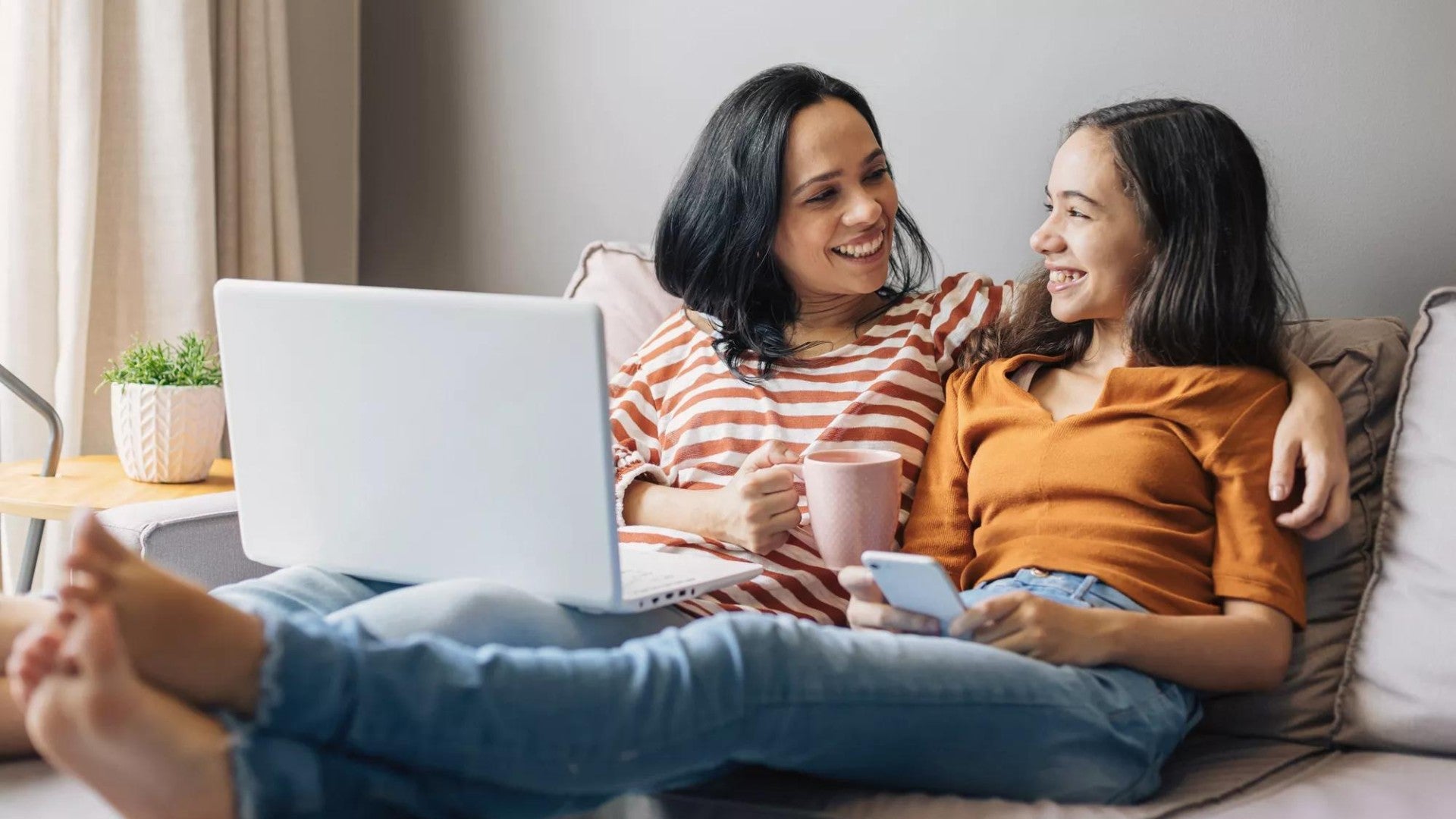Mother and daughter sitting together on a couch looking at a computer and talking about money.